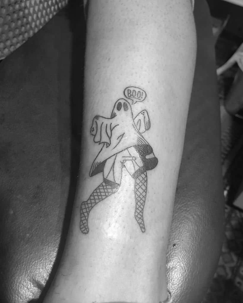 A black ink tattoo of a ghost wearing fishnet stockings and a jacket, lifting its sheet while saying 'BOO!' in a playful and humorous design.