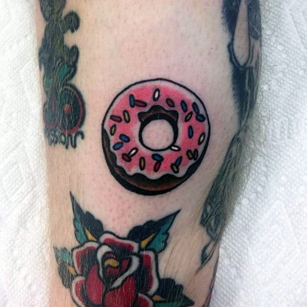 Small tattoo of a pink frosted donut with colorful sprinkles, outlined in black, placed among traditional-style tattoos.