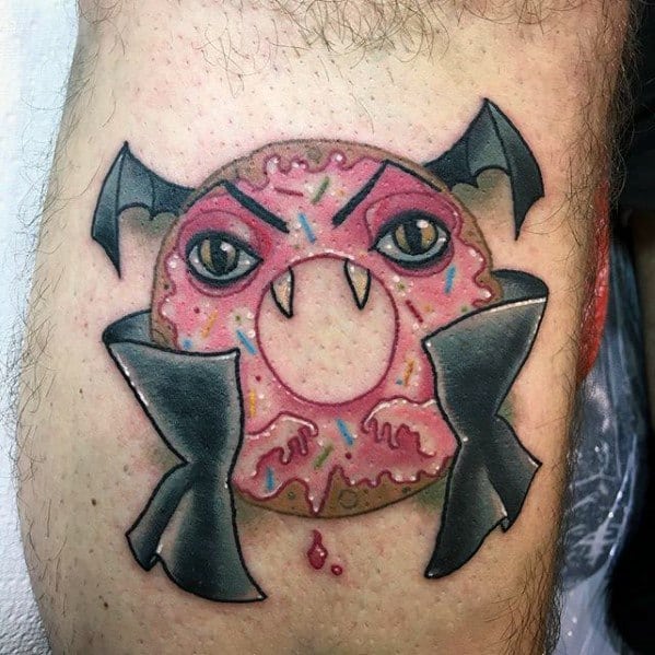 Tattoo of a pink frosted donut with vampire fangs, angry eyes, bat wings, and a black cape for a spooky yet fun look.