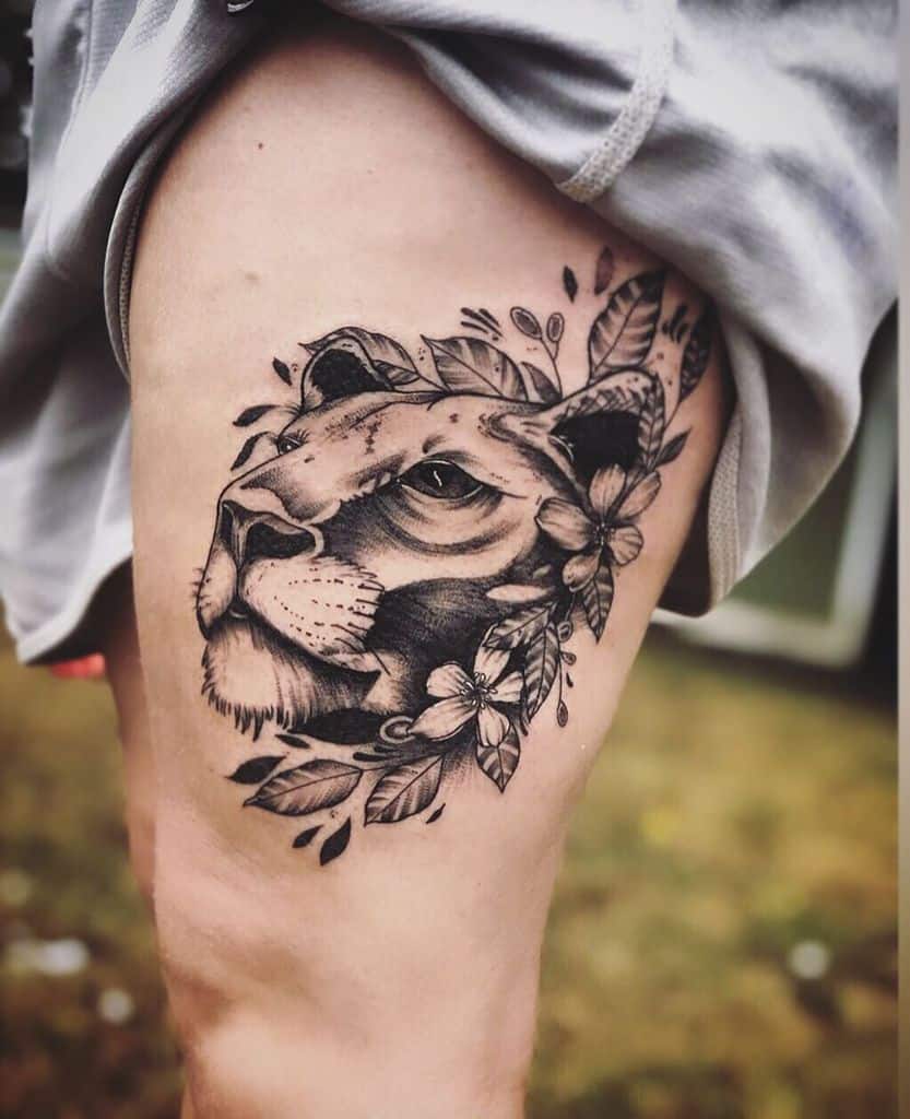 Black and grey lioness thigh tattoo with bold shading, floral accents, and detailed leaves, creating a fierce yet elegant design.