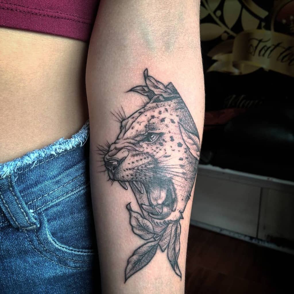 A striking black and grey tattoo of a roaring jaguar's face, surrounded by leaves, placed on the forearm for a fierce and detailed design.