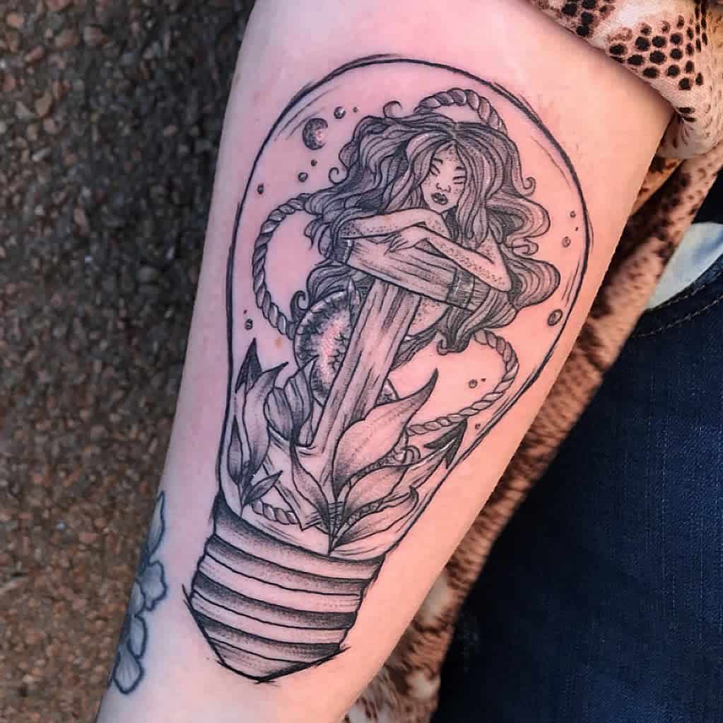 Black and grey tattoo of a mermaid wrapped around an anchor inside a light bulb, blending nautical and surreal elements with intricate details.