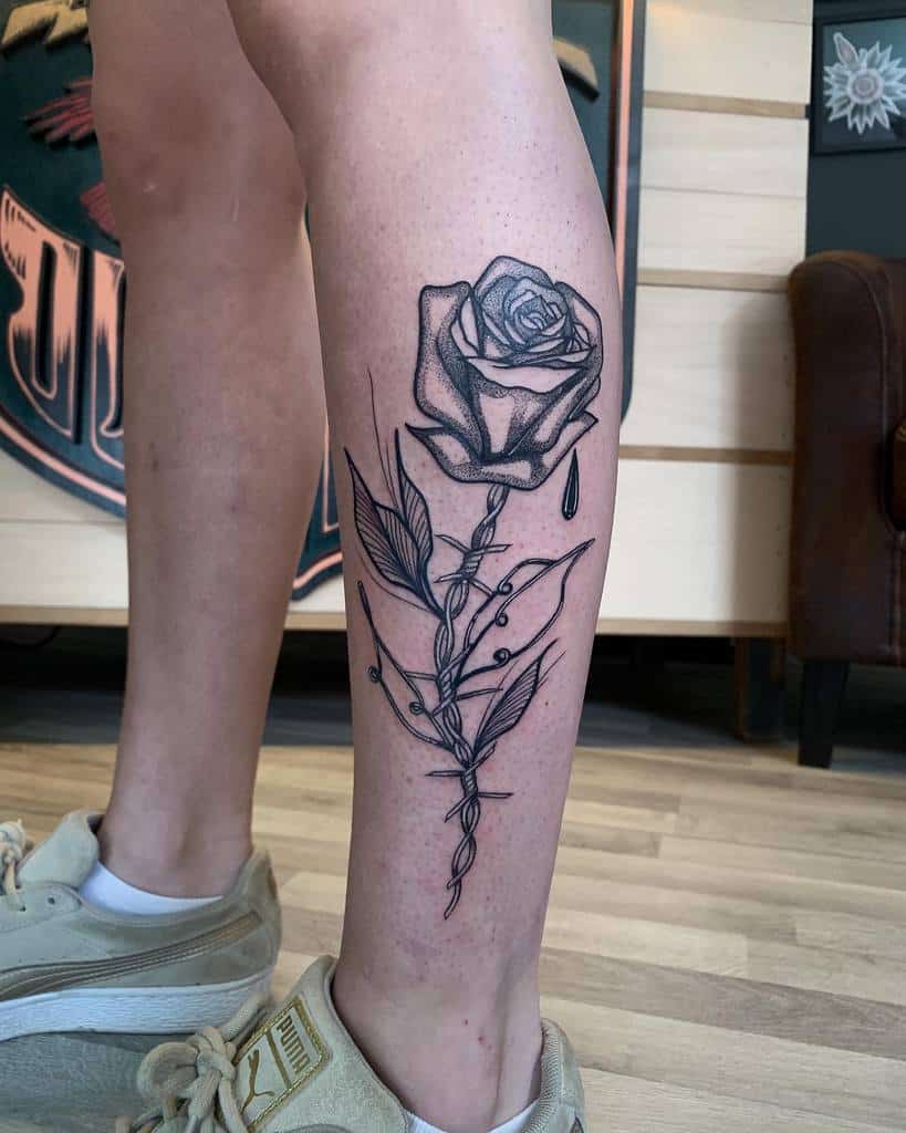 A detailed black and white tattoo of a rose with intricate shading and a delicate tear drop, placed on the calf with fine line work on the stem and leaves