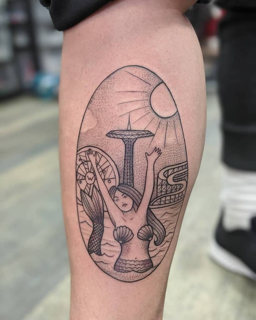 Black and grey tattoo of a mermaid with shell bikini, arms raised, in front of a cityscape featuring a Ferris wheel, bridge, and sun.