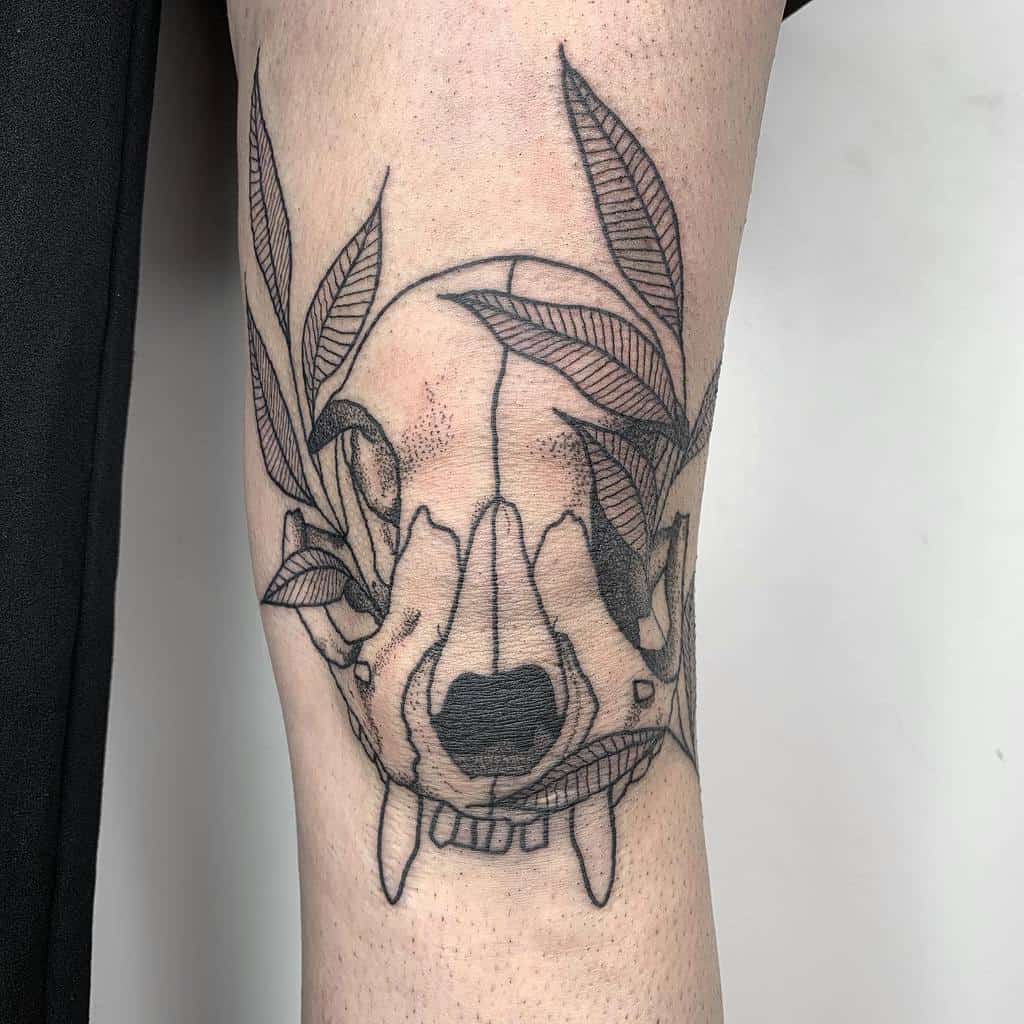 Tattoo of a cat skull adorned with leaves, located on the knee