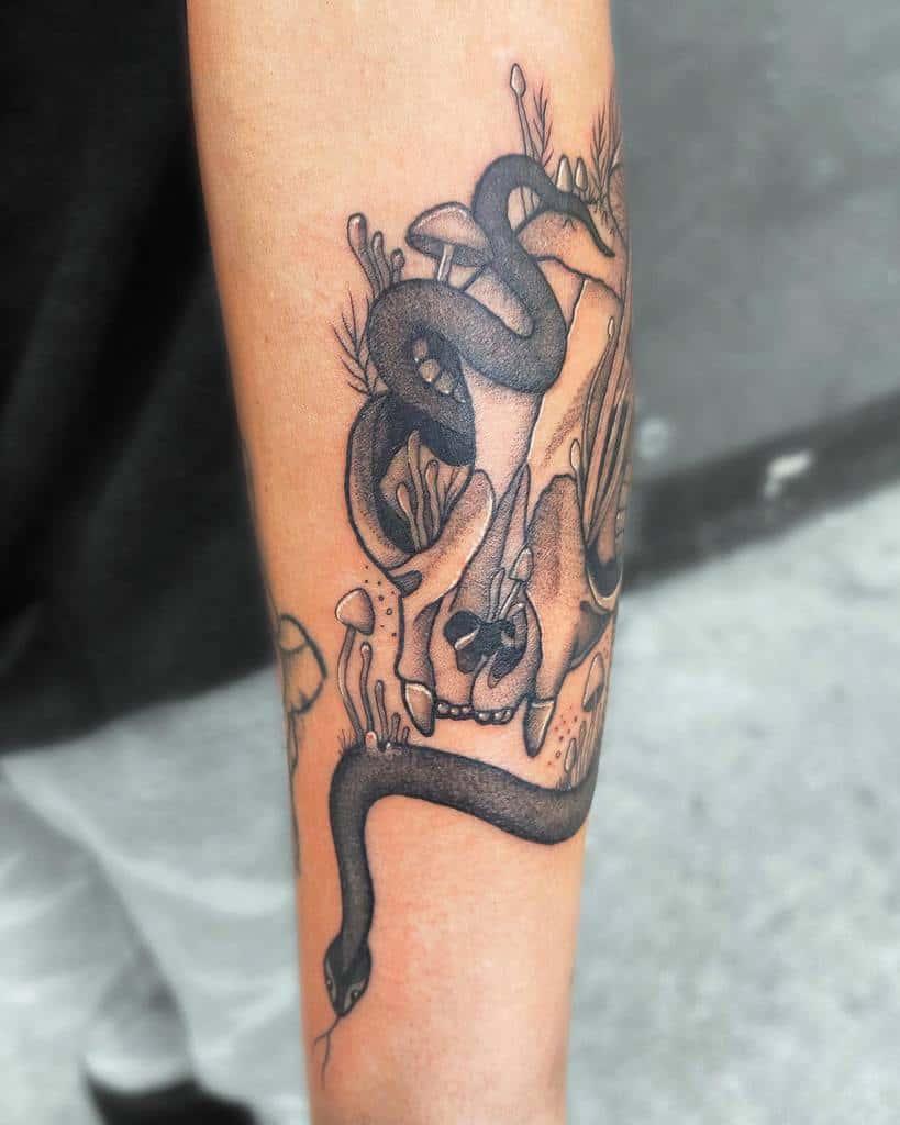 Forearm tattoo of a snake intertwined with mushrooms, plants, and a cat skull depicted in detailed black ink