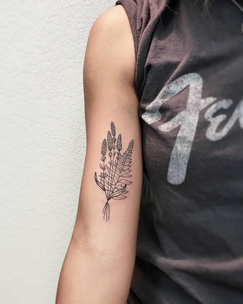 A black-and-white tattoo of a bunch of lavender and fern leaves arranged on the upper arm, featuring fine line details and delicate shading. The design has a simple, minimalistic aesthetic.