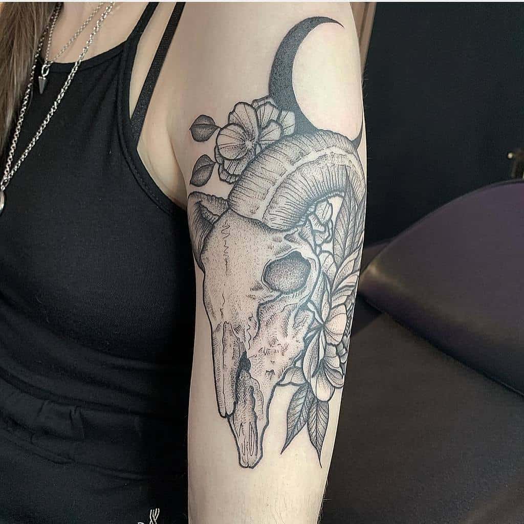 A detailed tattoo featuring a majestic ram skull adorned with flowers and a crescent moon elegantly graces an upper arm