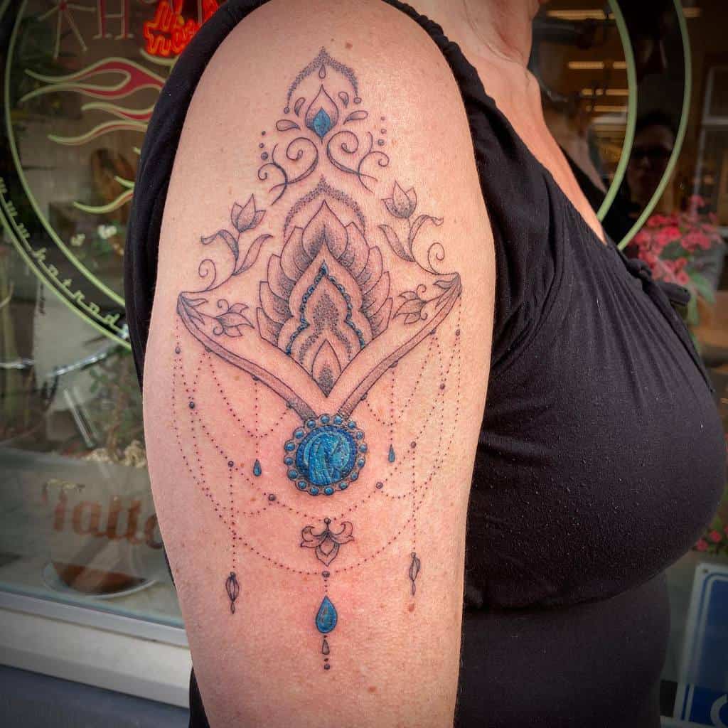 Ornate tattoo on arm with blue gem, floral patterns, and dangling beads