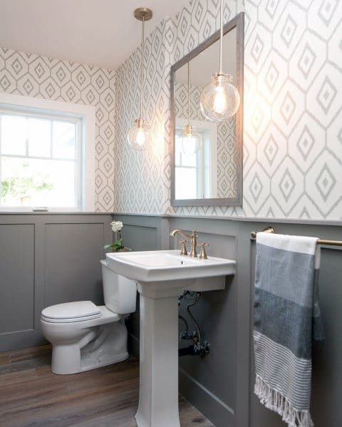 Bathroom with white patterned wallpaper, gray wainscoting, and hanging pendant lights.