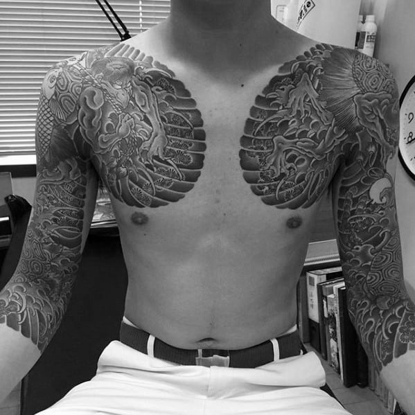 Shirtless man with detailed full-sleeve and chest tattoos sitting indoors