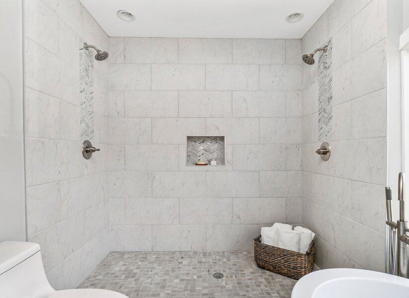 Spacious shower with two showerheads, tiled walls, a built-in niche, and a basket of towels on the floor
