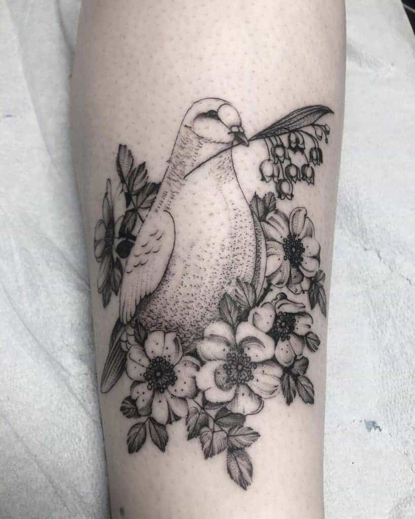 Detailed black and grey tattoo featuring a dove holding a sprig of lilies of the valley, surrounded by blooming flowers and leaves