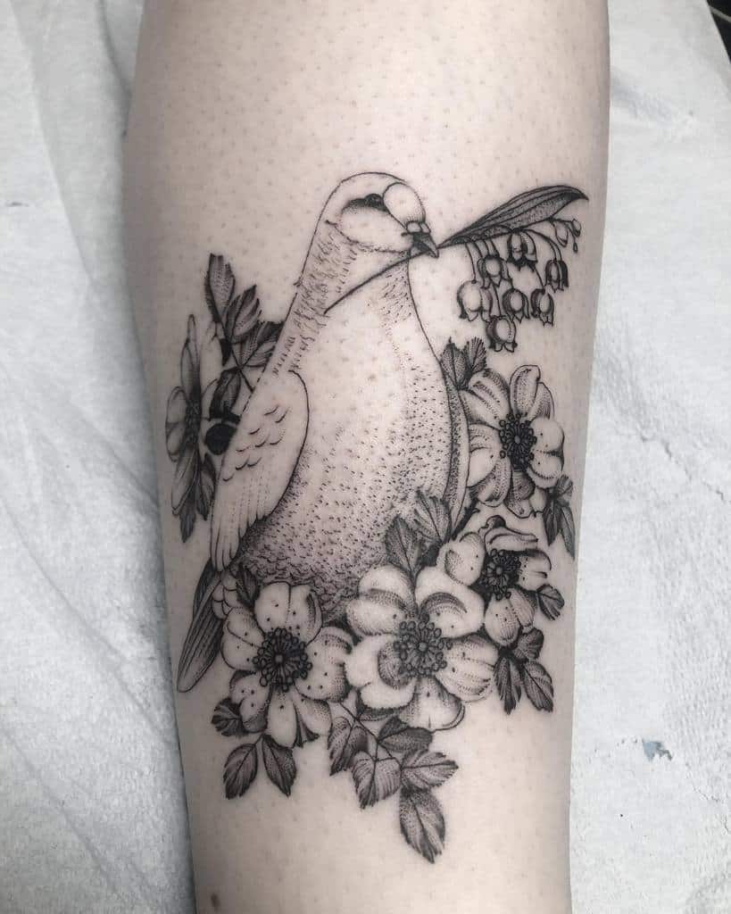 Black and grey tattoo of a bird holding a Lily of the Valley sprig, surrounded by detailed flowers and leaves, with fine dot work shading