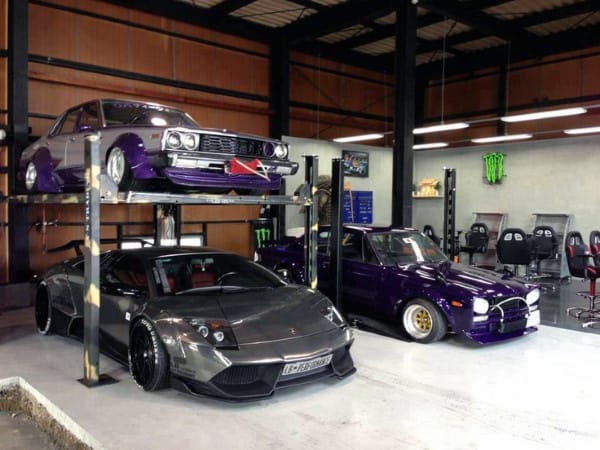 Three cars in a garage: a purple car on the lift, a gray sports car beneath it, and another purple beside them