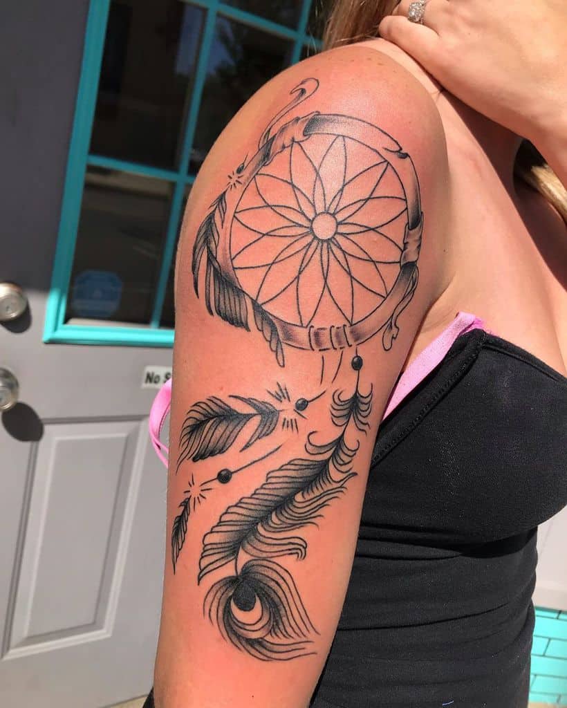 Black ink dreamcatcher tattoo with detailed peacock feather and floating beads on upper arm, featuring fine linework and shading.
