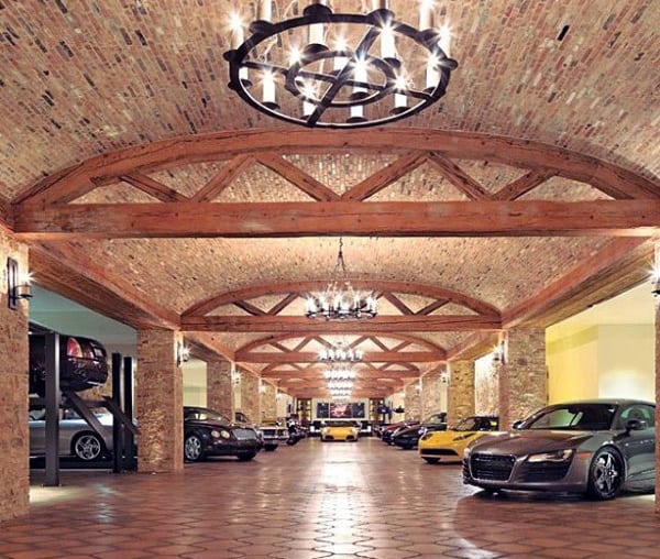 A luxurious garage with a rustic charm, featuring brick walls, arched timber beams, and elegant chandeliers, displaying an array of high-end cars.