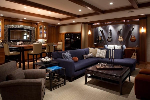 Stylish man cave with cozy blue sectional sofa, bar area, and guitar display wall under warm recessed lighting
