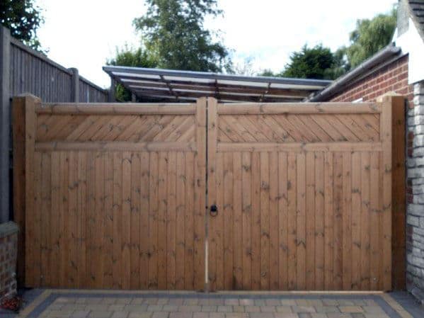 Wooden double gates with diagonal panel design, set on a brick driveway, with trees and a tall fence in the background
