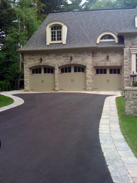 Asphalt driveway with stone edging leading to a stone house with three arched garage doors
