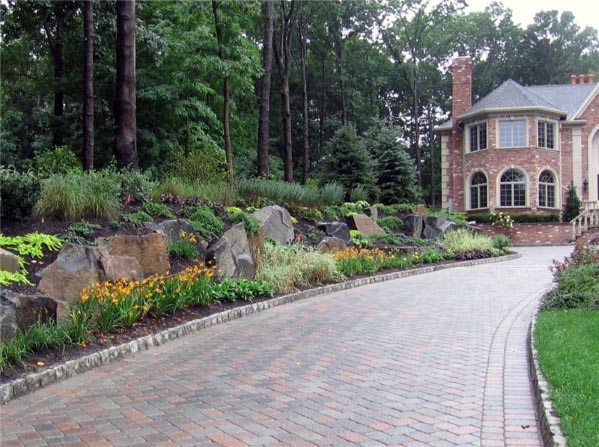 Brick-paved driveway with stone edging bordered by landscaped rocks and vibrant flowers.