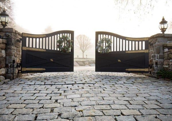 Black wooden driveway gate with arched top, brass accents, and festive wreath decorations.