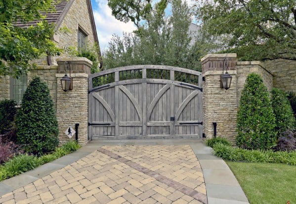 Rustic wooden arched gate with natural finish, framed by stone pillars, complementing a traditional house setting.