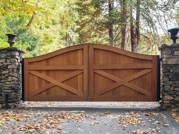 Natural wood driveway gate with arched top and X-brace design flanked by stone pillars.