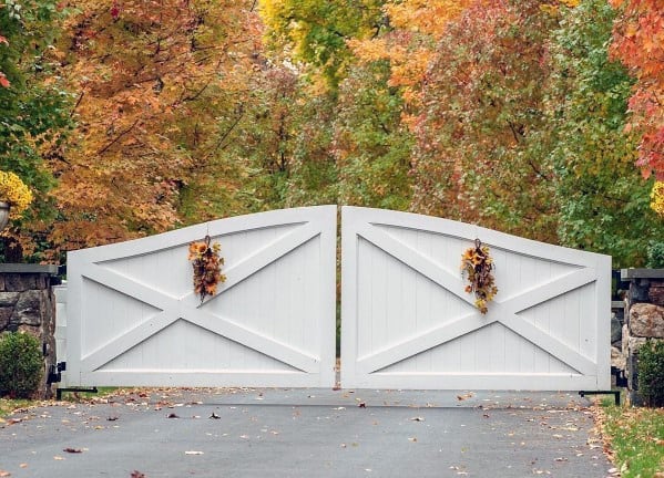 White painted wooden double gate with cross-brace design, adorned with autumn wreaths, blending seamlessly with the fall foliage.