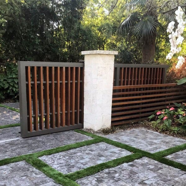 Wooden slat gate with white stone pillar, surrounded by greenery and a tiled pathway
