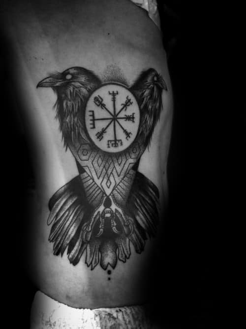 Tattoo on the side of a torso featuring two ravens and a circular Nordic symbol