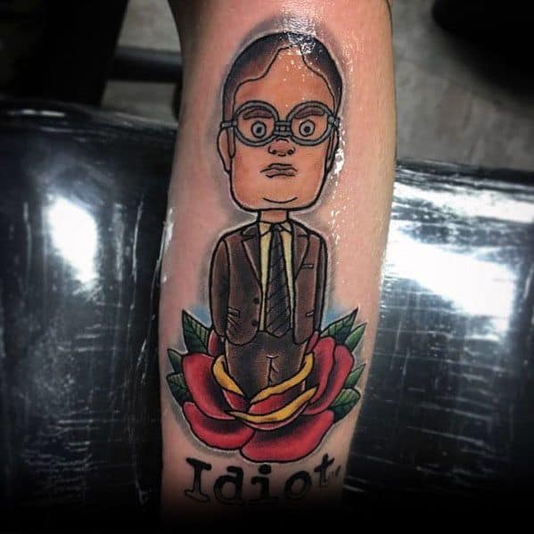 Cartoon-style tattoo of Dwight Schrute with an exaggerated head, standing in a red and yellow rose, with the word "Idiot" written below.