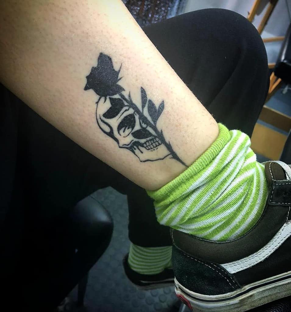 Minimalist black ink tattoo of a skull with a rose, featuring bold contrast and clean lines on the lower leg.