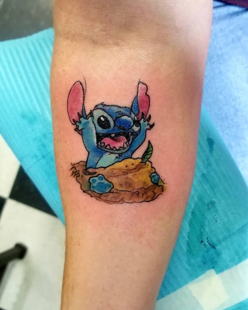 Colorful tattoo of Stitch from Lilo & Stitch emerging from a cookie with a playful expression, on the lower arm.