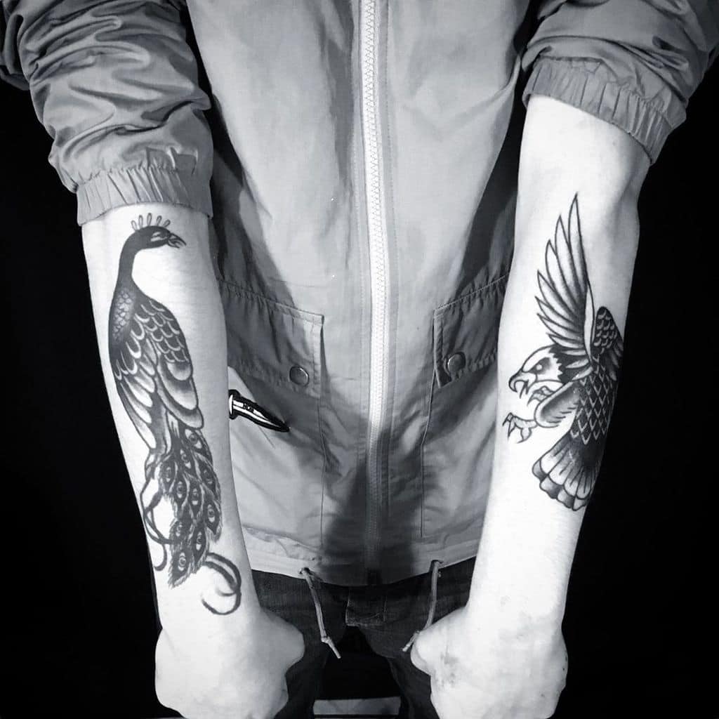 Black and white tattoos of a peacock on one arm and an eagle on the other, featuring detailed feather patterns.