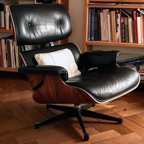 Black leather Eames lounge chair with wooden frame in a cozy library setting.