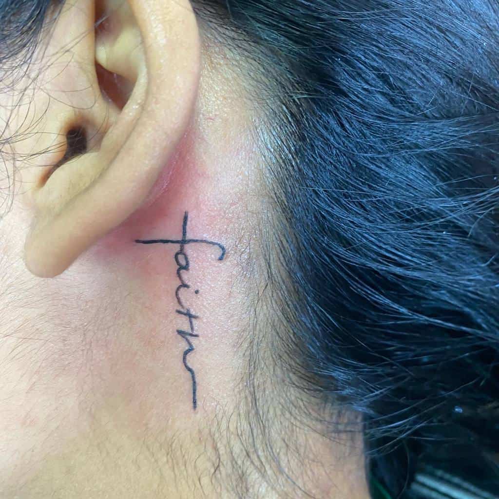A delicate tattoo of the word "faith," elegantly scripted in cursive, graces the area near a person's ear, with a tiny cross subtly intertwined within the design