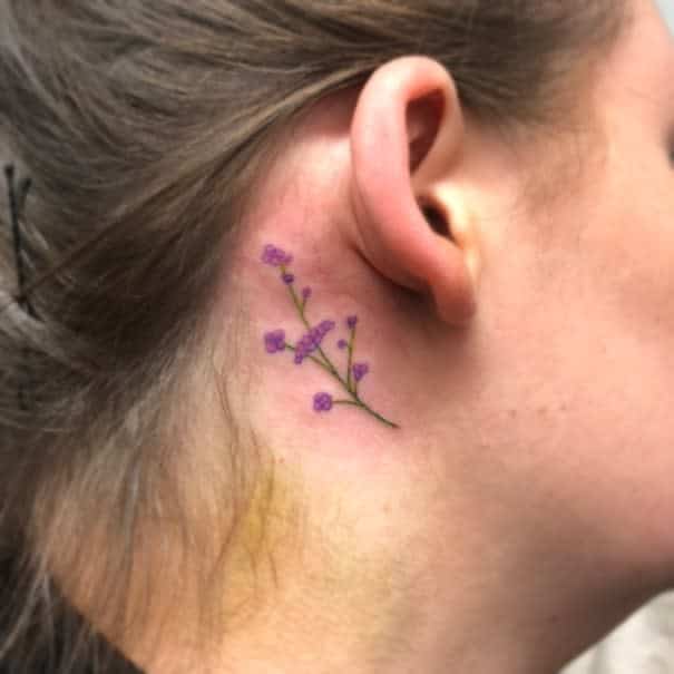 Minimalist behind-the-ear tattoo of a delicate violet sprig with tiny purple flowers, symbolizing grace, femininity, and elegance
