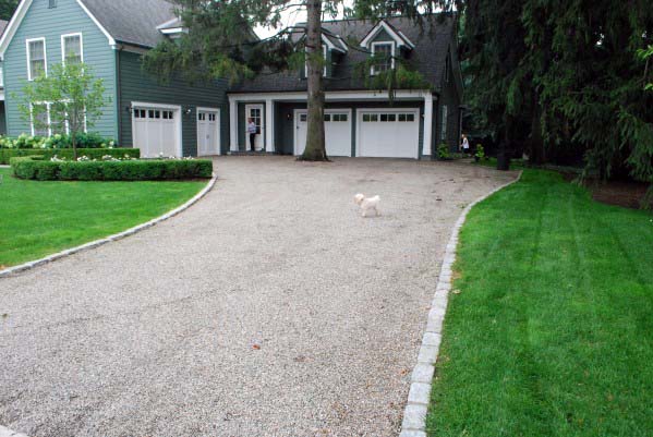 Gravel driveway with stone edging leading to a green house with a three-car garage and landscaped lawn.