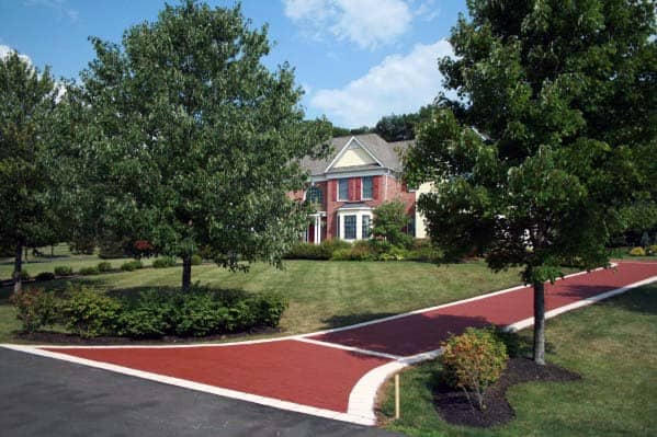 Red brick driveway with white concrete edging leading to a brick house surrounded by trees.