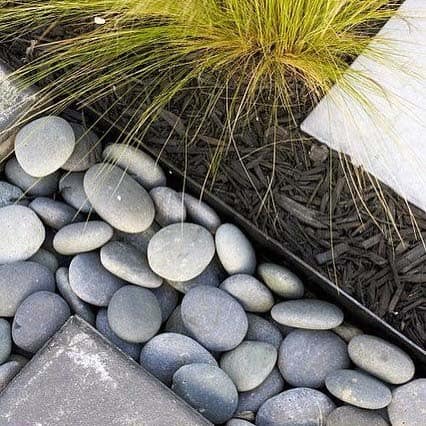 Luxury river rock landscaping with smooth dark pebbles, bordered by modern stone tiles and lush greenery