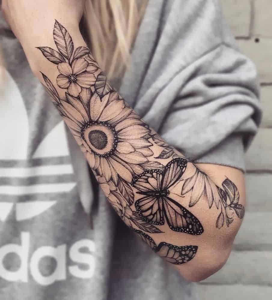 Black and grey full forearm tattoo featuring a detailed sunflower, smaller flowers, leaves, and butterflies with intricate shading and fine linework.
