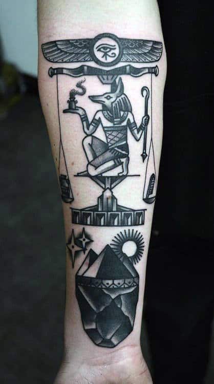 Black and grey tattoo of Anubis with scales and a mountain landscape on the forearm