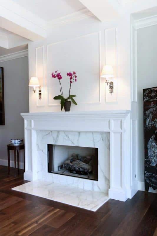 White marble fireplace with a classic mantel, flanked by wall sconces, and adorned with orchids.