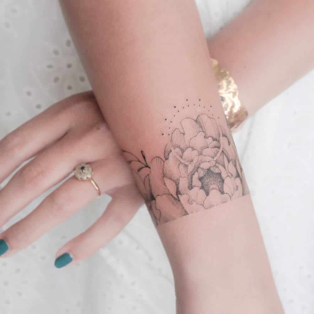 Elegant black ink floral tattoo with soft shading, wrapping around the wrist, featuring a large blooming flower