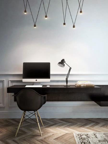 Minimalist workspace with a black chair, desk, computer, lamp, and books; suspended lights above herringbone floor