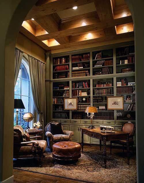 Cozy library featuring a high bookshelf, comfy armchair, leather ottoman, and wooden desk bathed in warm lighting