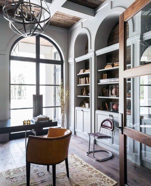 Elegant home office with arched shelves, large windows, leather chair, and rustic chandelier.