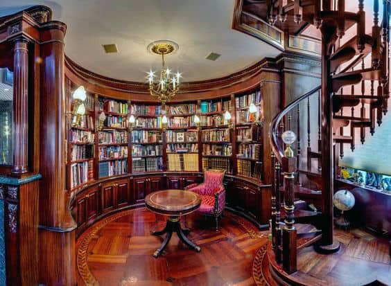 A round wooden library with a red chair, spiral staircase, chandelier, and shelves brimming with books