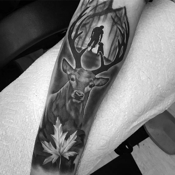 Realistic black and grey elk tattoo on the forearm, featuring a forest scene within its antlers and a maple leaf for a symbolic, nature-inspired design.